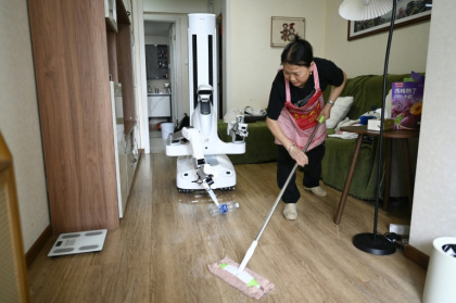 Un robot assistant ménager transporte une bouteille jusqu'à une poubelle tandis qu'une femme de ménage nettoie le sol chez une cliente à Pékin, le 21 mai 2026 en Chine - Michael ZHANG, QIAN Ye (AFP)